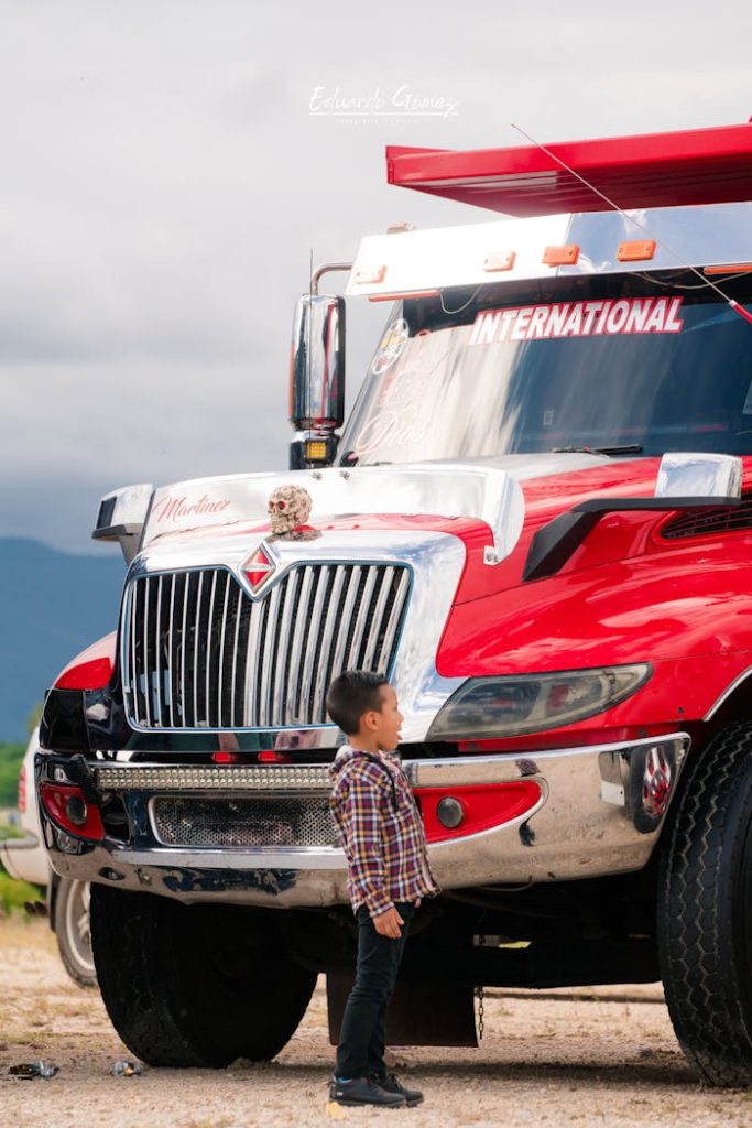 boy-standing-by-a-large-red-truck-17824895 A young boy stands captivated by a large red semi-truck parked outdoors, showcasing vibrant contrasts.